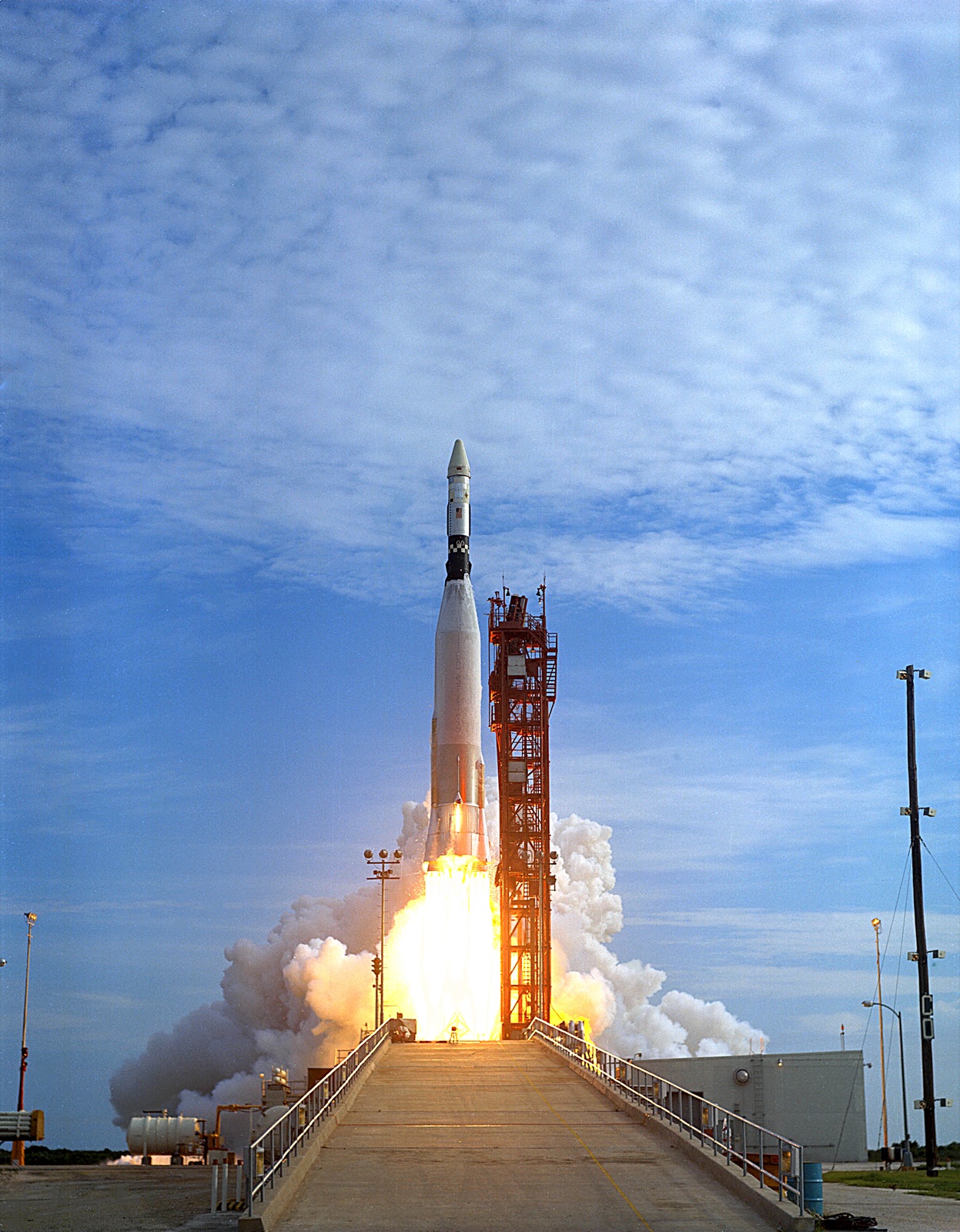 A NASA Atlas Agena rocket lifting off from its launch pad in a cloud of exhaust under a blue sky.