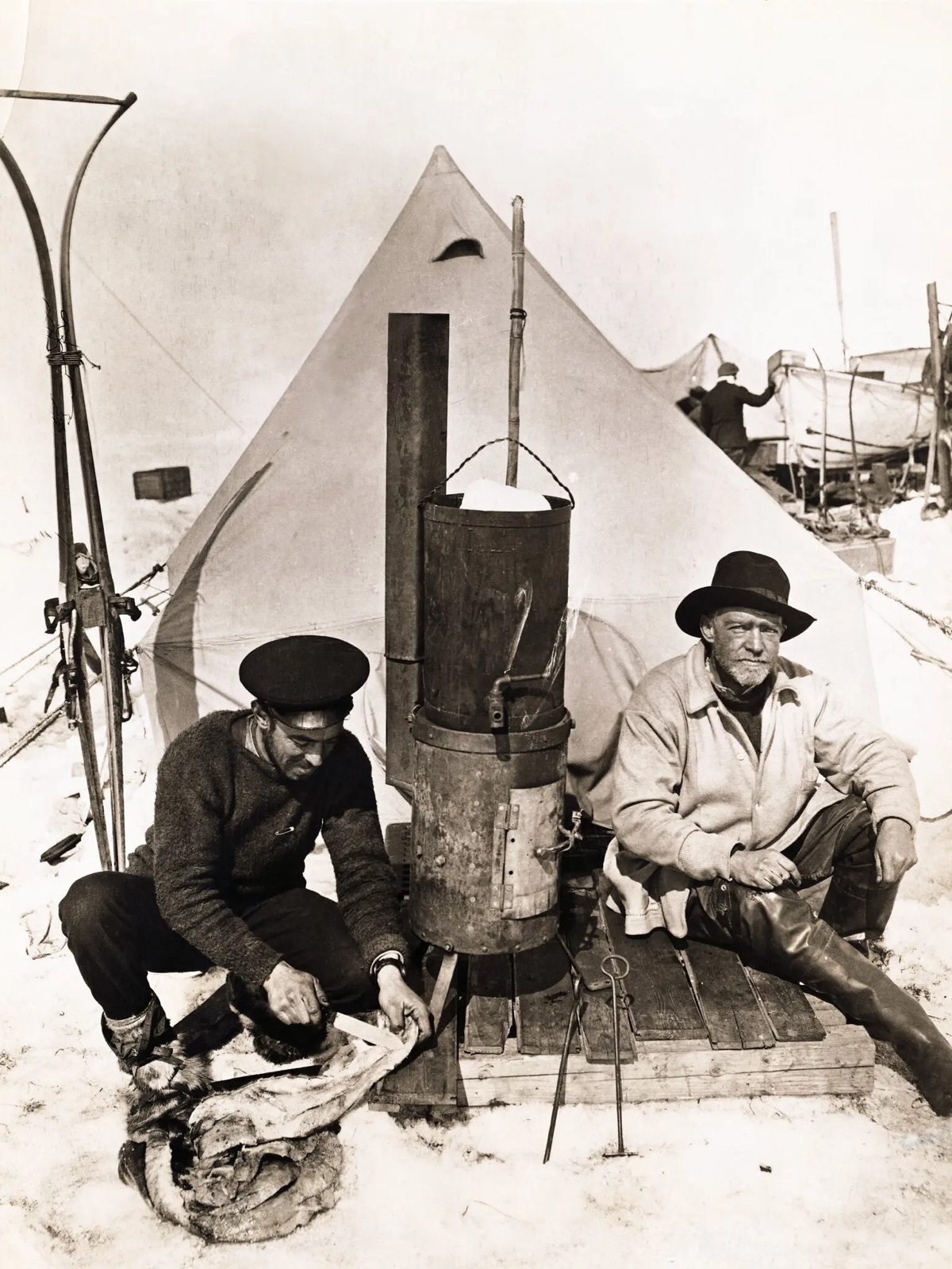 Two men of the Shackleton Antarctic expedition sitting beside a stove outside a tent on the ice, one of them Ernest Shackleton in a wide-brimmed hat, with a third figure at a boat in the background.