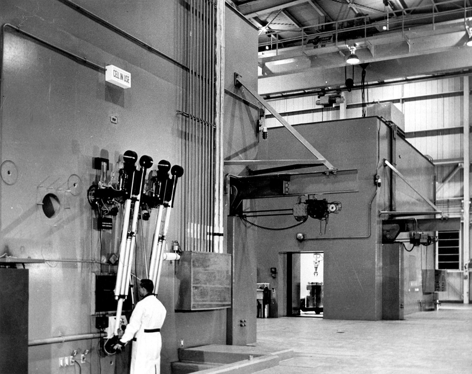 A technician in a white lab coat stands at the shielded wall of a hot cell, gripping the handles of master-slave remote manipulator arms beneath a lit sign reading CELL IN USE.