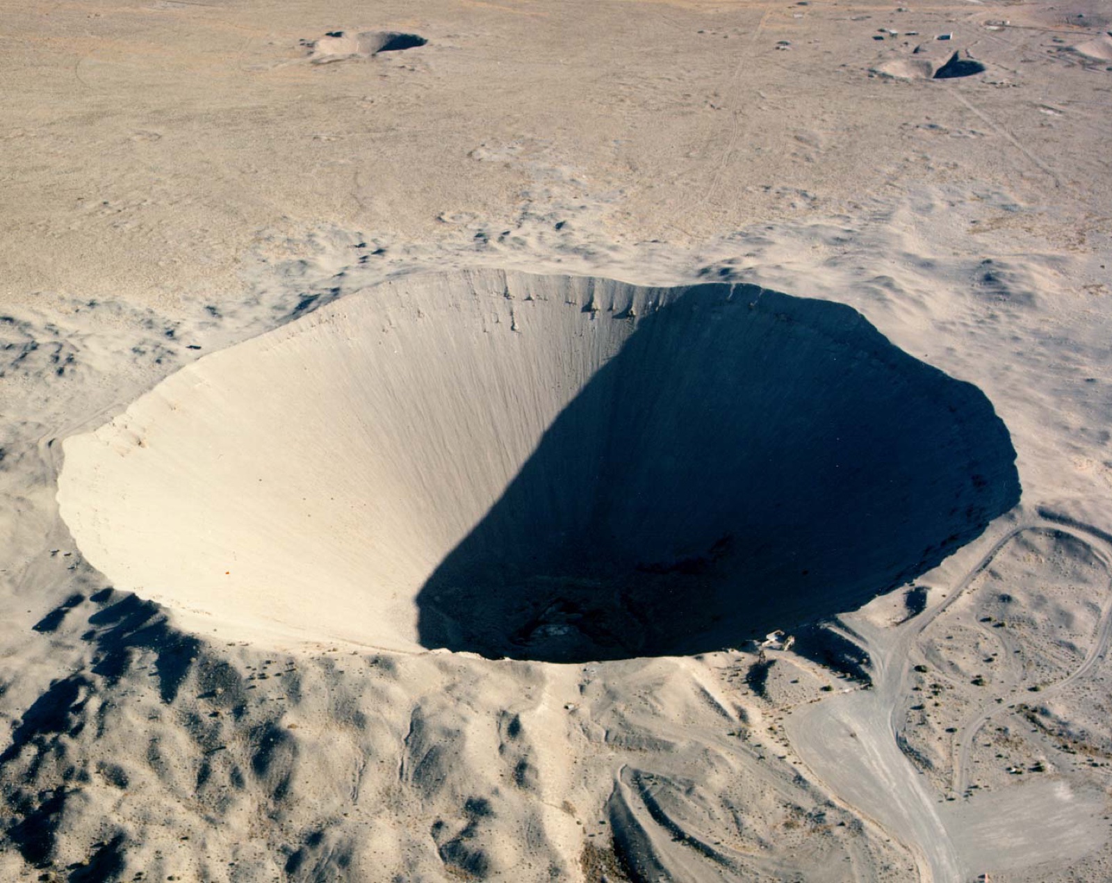 An aerial photograph of the Sedan crater at the Nevada Test Site — a wide, pale bowl of blasted desert, half in shadow, thrown up by a buried nuclear explosion in 1962.