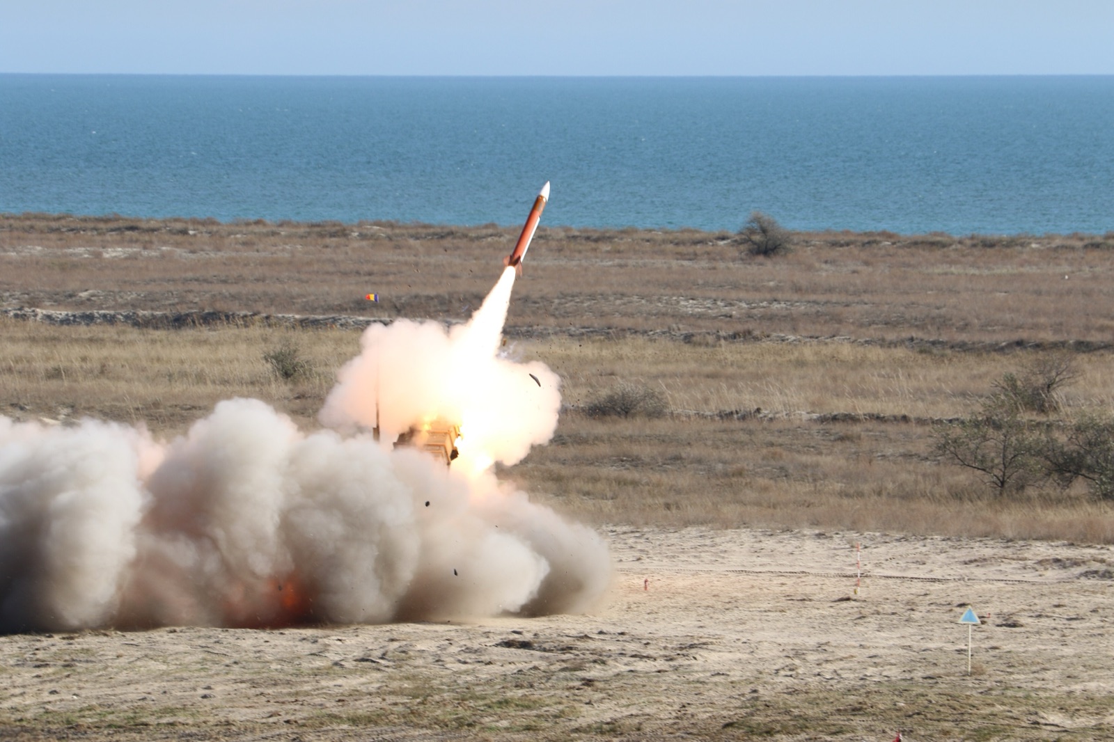 A Patriot air-defense missile lifting off its launcher in a cloud of exhaust on flat coastal scrubland, photographed during a live-fire exercise by the 74th Patriot Regiment of the Romanian Army.